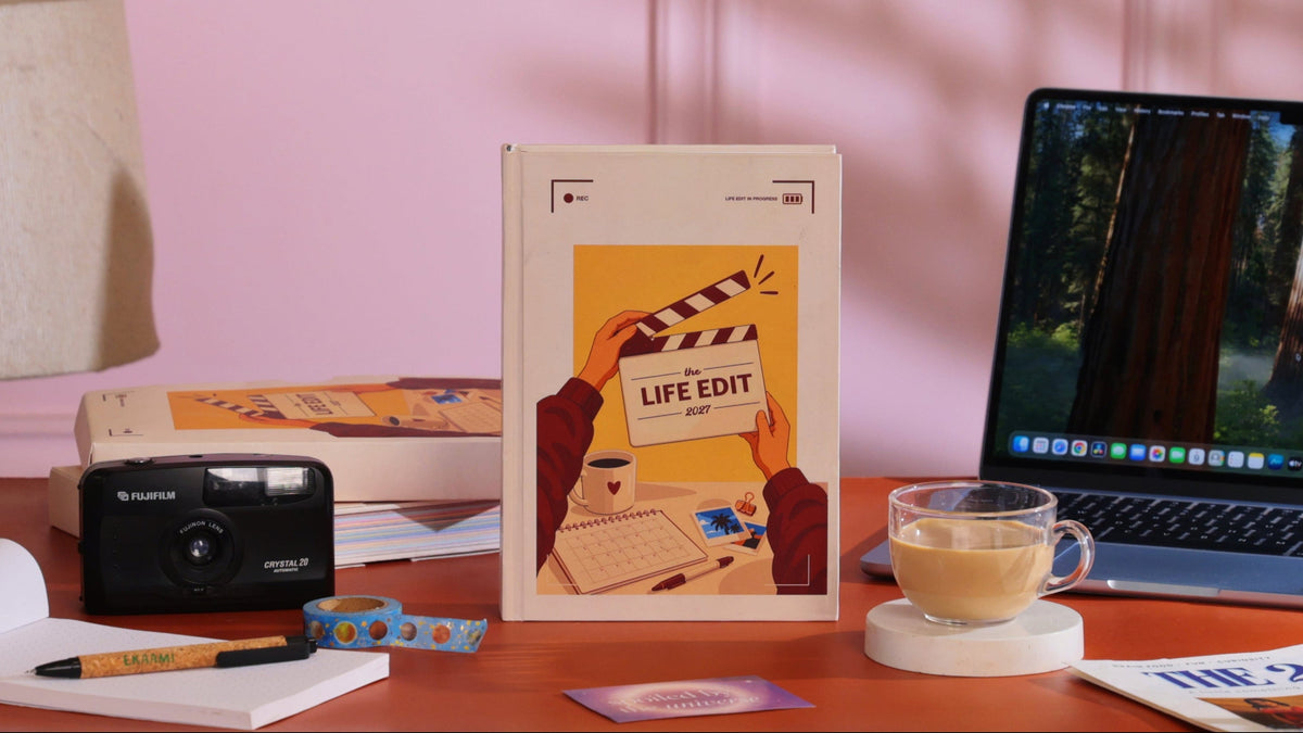 A book titled 'LIFE EDIT' with a warm yellow cover illustration stands on a wooden desk in a styled room setting. The scene includes a laptop, a glass mug of coffee, a notebook, a camera, and other desk accessories against a pink paneled wall with leaf shadows.